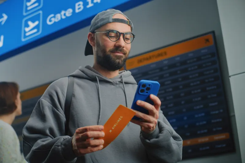 Reisender mit Bordkarte und Handy schaut vor der Abflugtafel auf sein Display